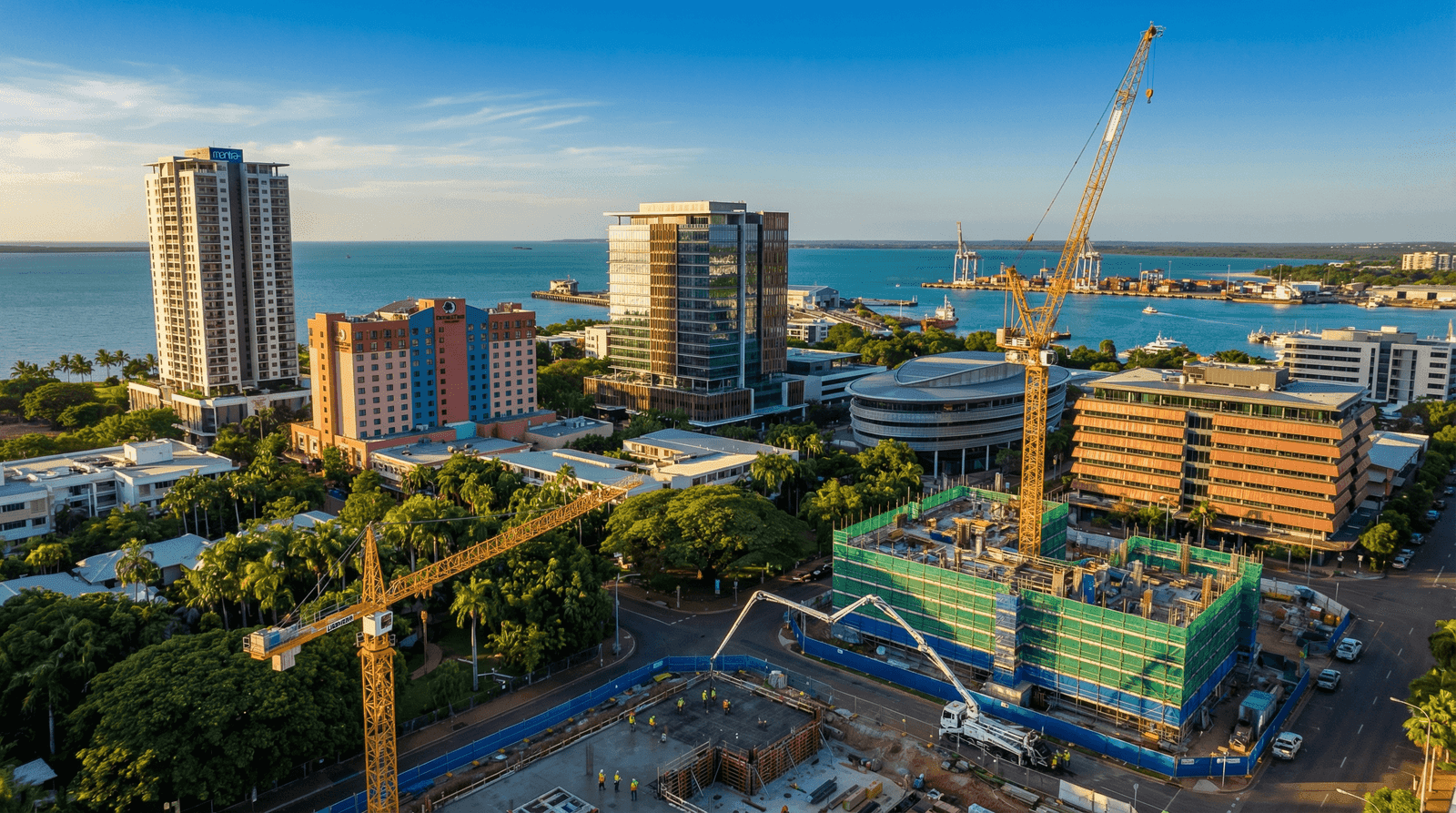 Aerial view of Darwin CBD with yellow Liebherr tower cranes, multi-storey construction sites wrapped in green scaffold mesh, tropical tree canopy, and turquoise Darwin Harbour with port facilities in the background